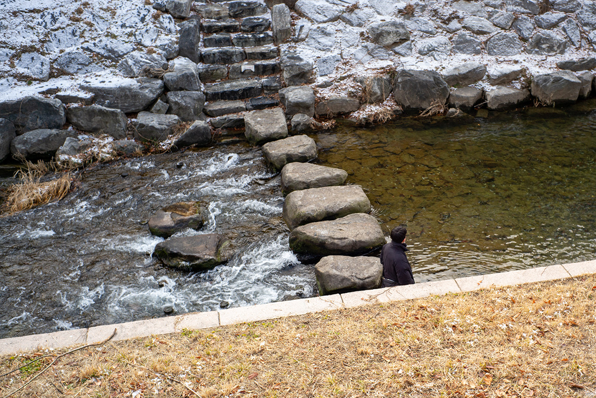 겨울의 개천과 돌다리 위를 흐르는 물, 강가에 앉아 있는 인물의 뒷모습을 라이카 M-P Typ 240 특유의 차분한 색감으로 담은 거리 사진