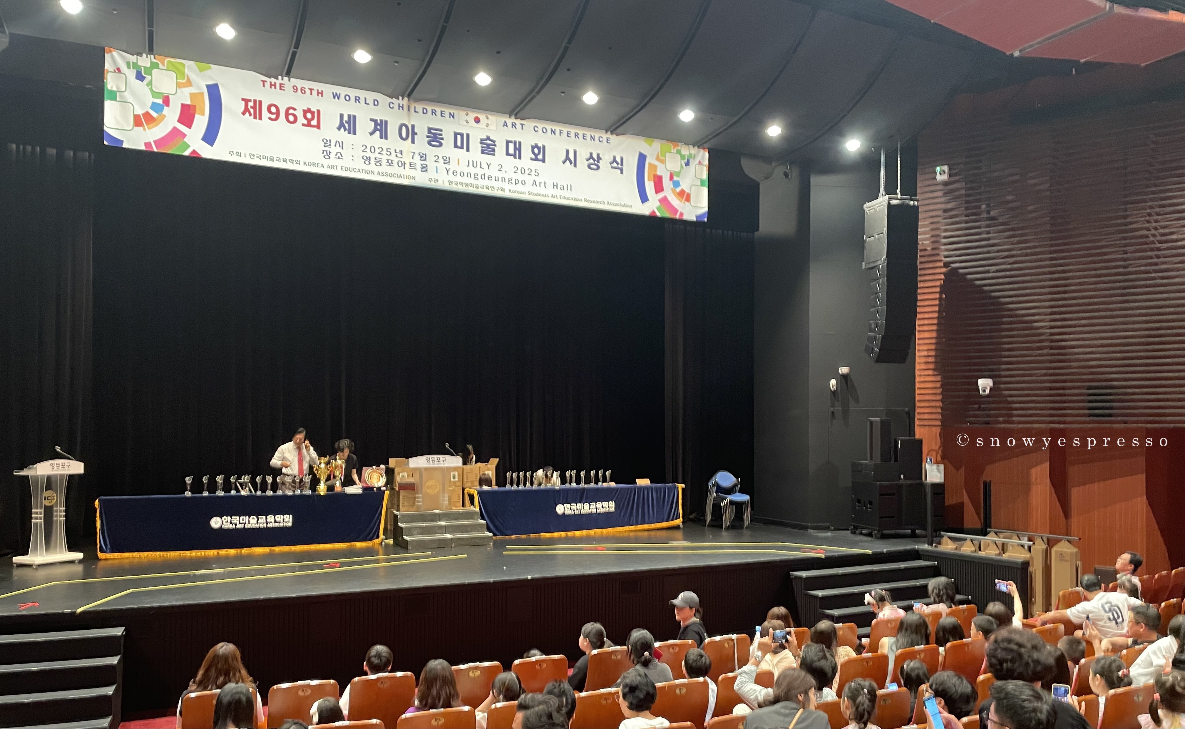 재96회 세계아동미술대회 시상식 단상에서 관계자들이 사전 준비하는 모습과 객석에 앉은 수상자들