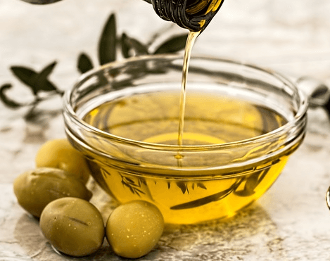 투명 유리 그릇에 노르스름한 올리브유가 가득하고, 그릇 앞에는 연두색의 올리브 4개가 있다.