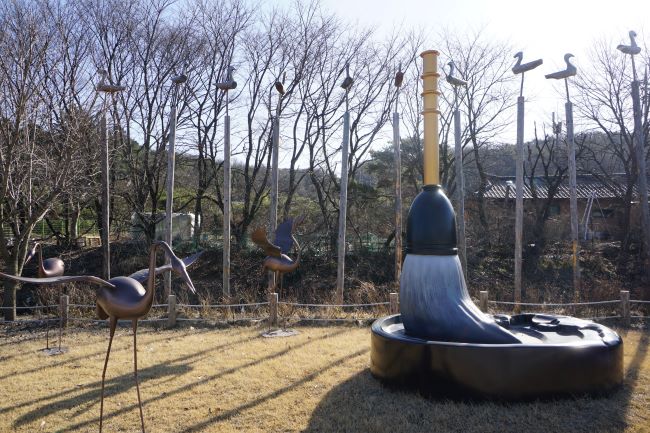 큰 벼루 붓 조형물, 그 뒤로 많은 솟대들, 옆에는 두루미 조형물,