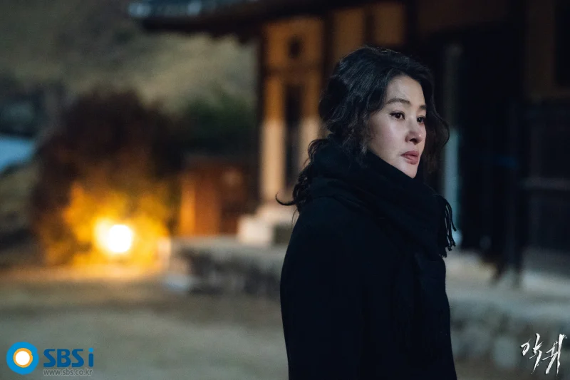 어느 한 시골 전통 가옥을 배경으로 검은색 롱코트를 입고 어딘가를 응시하고 있는 윤경문