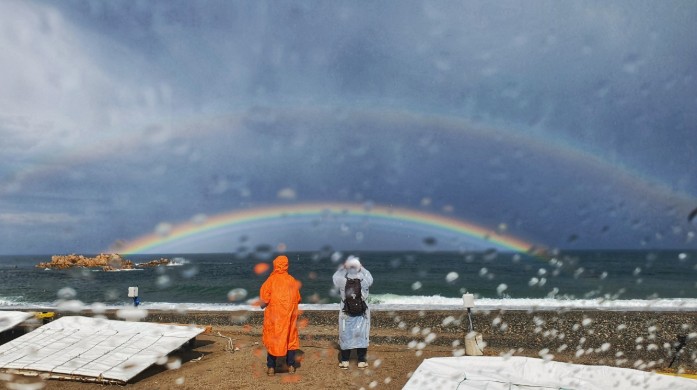 Two hikers standing in the rain watching a rainbow over the sea — somewhere on the Korean coast