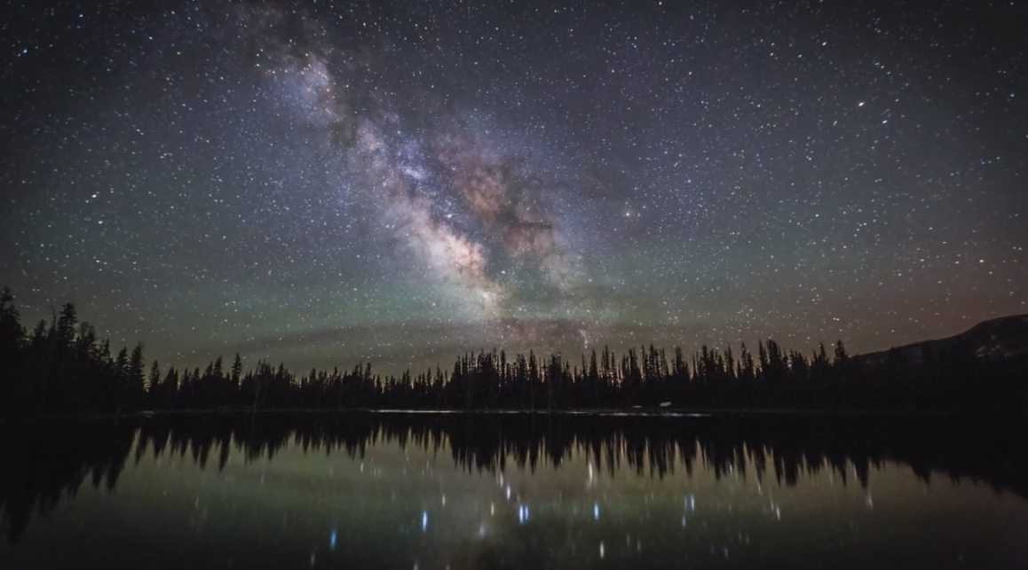 "밤하늘에 펼쳐진 은하수와 별들이 고요한 호수 위에 반사되는 장면, 주변에는 침엽수림이 어두운 실루엣으로 둘러싸여 있음"
