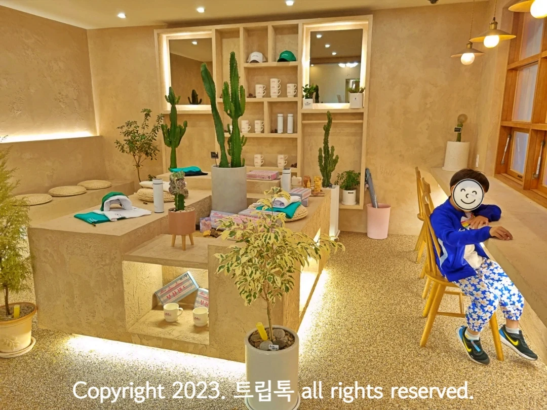 베이색 내부에 가운데 화분과 소품이 있고 오른쪽에 창문쪽의 의자에 한아이가 앉아있다. 