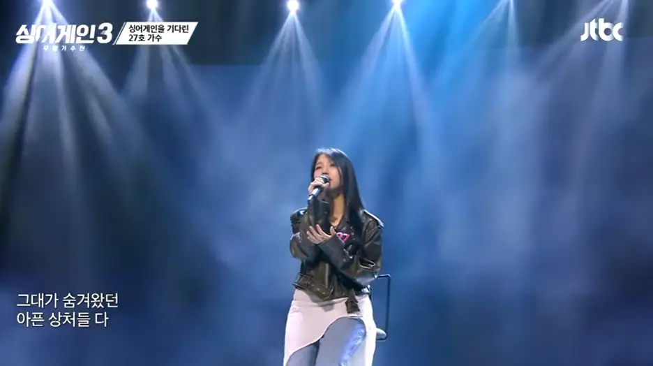싱어게인3- 긴생머리에 오른손으로 마이크를 쥐고 청바지 위에 흰색 치마를 두른채 슬프게 노래하는 27호