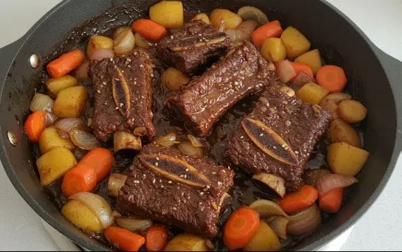윤기 나는 갈비찜과 감자, 당근, 양파가 팬에서 조리되고 있는 모습