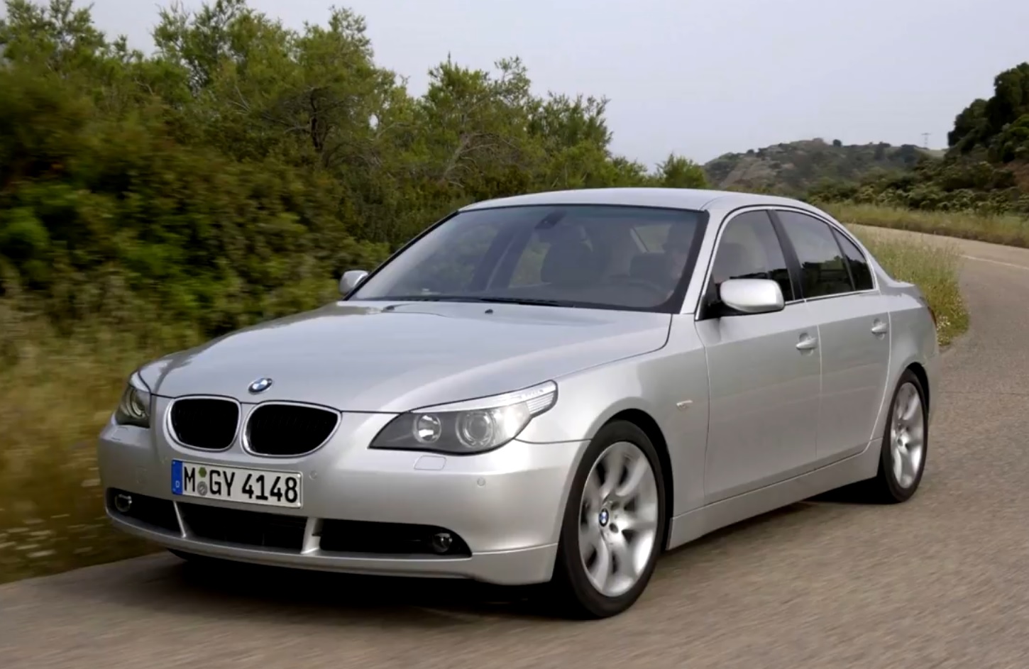 A dynamic rolling shot of a dark grey BMW 5 Series E60 driving fast on a winding forest road, showcasing its sharp eagle-eye headlights and unique sculpted body lines