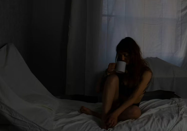 커튼 사이로 희미한 새벽 빛이 들어오는 방에서, 침대에 앉아 머그잔을 들고 조용히 아침을 준비하는 여성