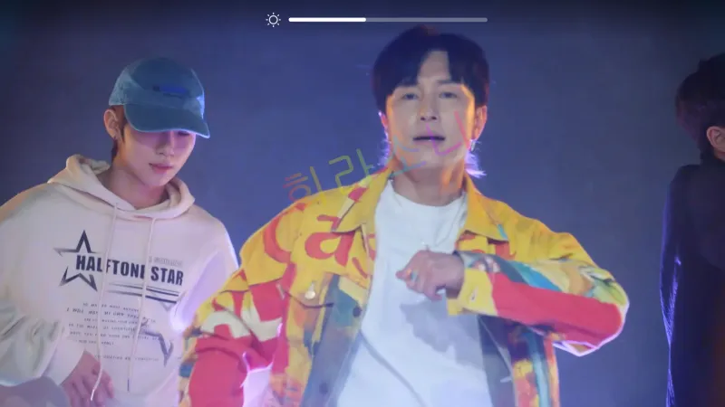 무대 위에서 오흥수(김동완)가 춤을 추고 있다. 오흥수는 흰색 티셔츠 위에 알록달록한 노란색 재킷을 걸치고 있으며&amp;#44; 박력 있는 표정으로 춤을 추고 있다. 그의 왼쪽에 있는 남성은 베이지색 후드티와 파란색 야구 모자를 착용하고 있으며&amp;#44; 조용히 춤을 맞추고 있다. 배경은 어두운 조명으로 연출되어 있다.