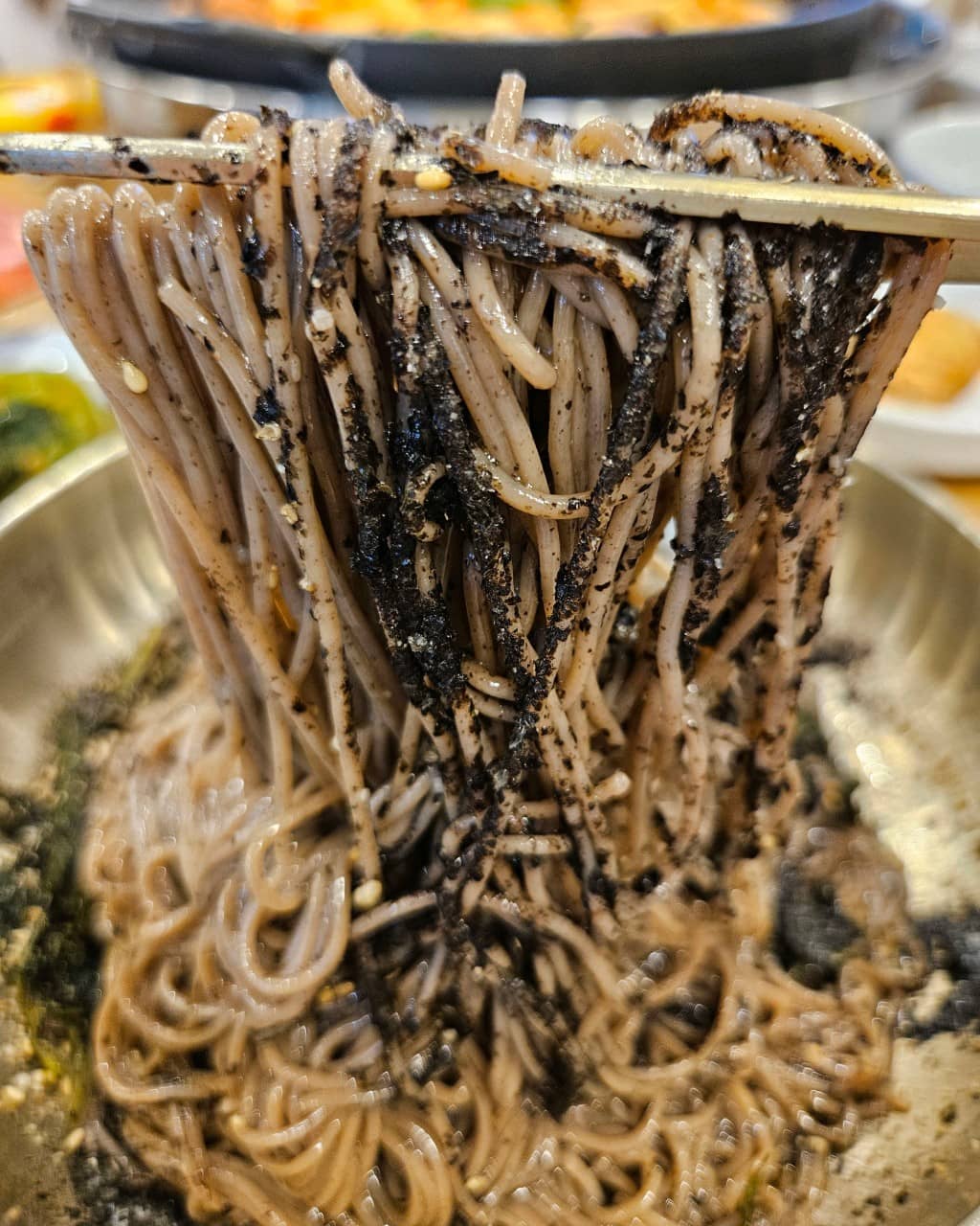 '개수리막국수' 들기름 막국수 사진입니다. 젓가락으로 막국수를 들어 올리고 있고 면에는 깨와 김가루가 뿌려져 있습니다.