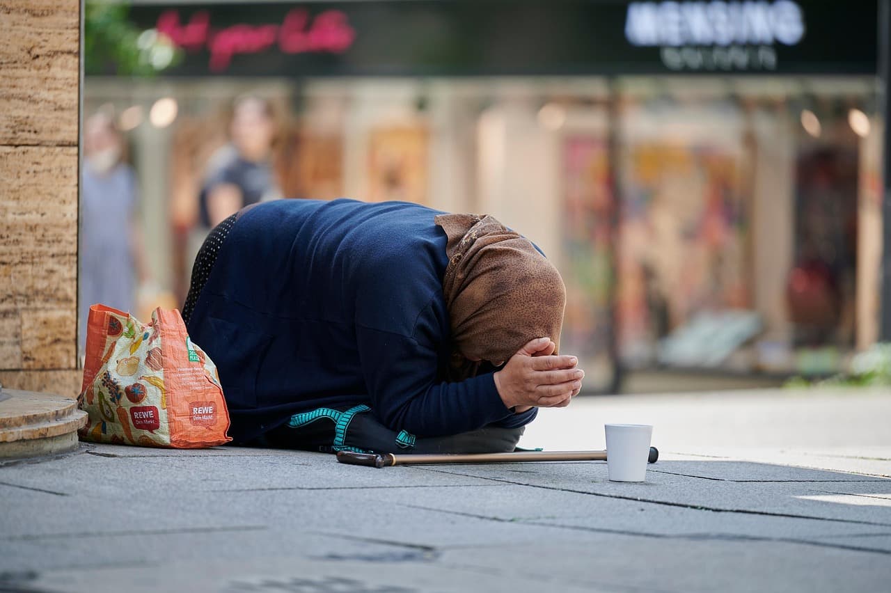 도심 땅바닥에 몸을 접은 채로, 손바닥을 맞대고 엎드린 여성 앞에 컵이 놓여 있어 구걸을 하고 있는 이미지로 빈곤한 저소득층을 상징하는 이미지로 연결