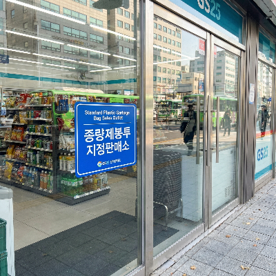 서울의 현대적인 편의점 유리문에 붙어 있는 파란색 '종량제봉투 지정판매소' 공식 인증 스티커. 깨끗한 도시 거리 배경과 낮 시간의 자연스러운 채광이 어우러진 8k 초고해상도 사진으로, 구매 가능 여부를 확인하는 가장 쉬운 방법을 시각적으로 설명함