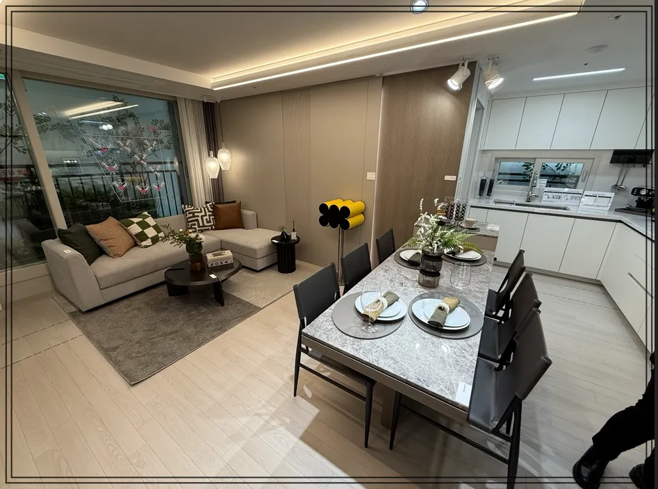 아파트-모델하우스-내부-오른쪽에-주방-싱크대와-6인-식탁이-있고-식탁위에-접시와-꽃병이-데코되어-있음-왼쪽으로-거실이-있는데-연그레이-쇼파와-검은색-테이블-쇼파위에-쇼파쿠션-5개-있음