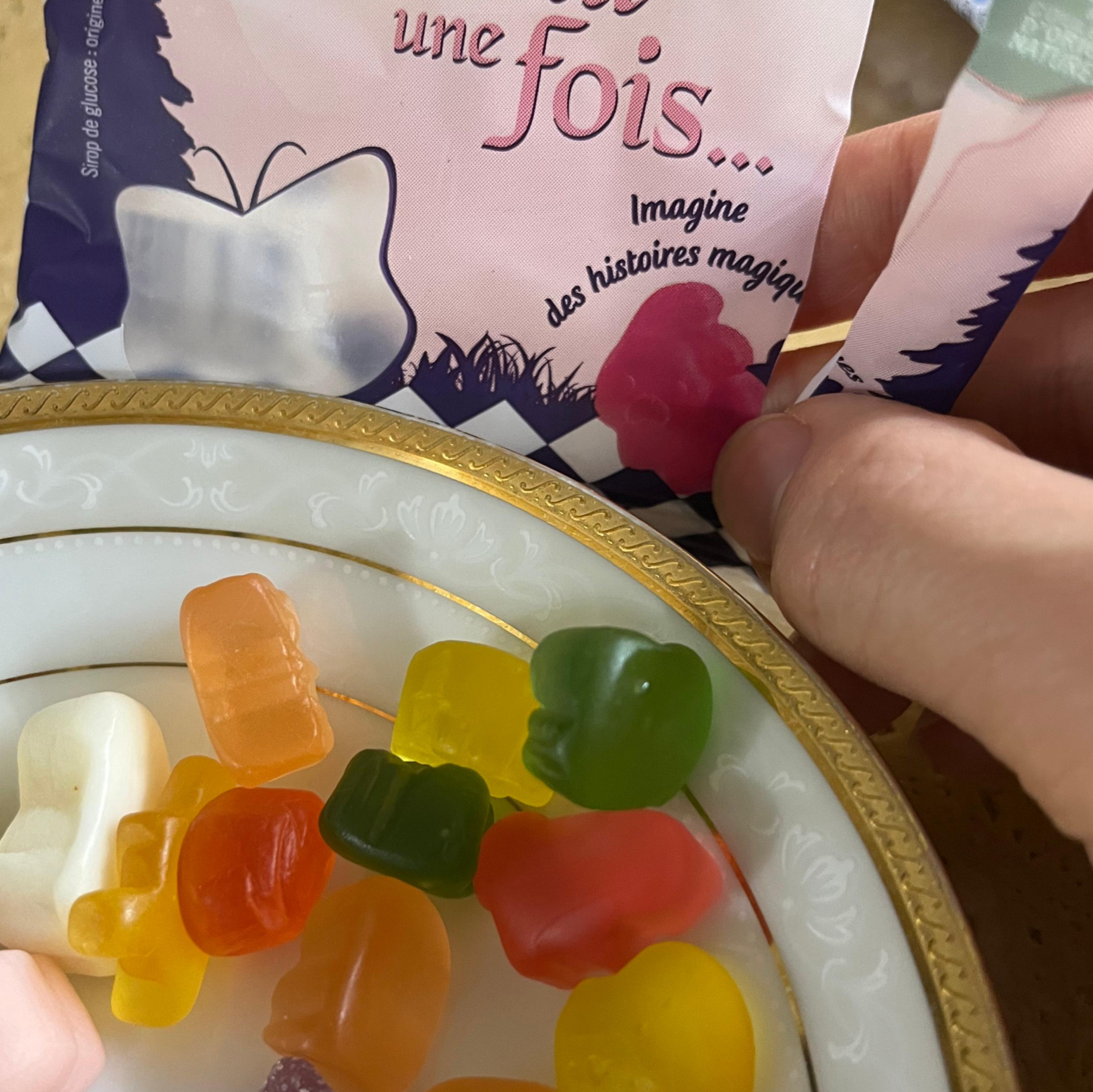 나비와 핑크빛 숲이 그려진 '원스 어폰 어 타임(Il était une fois...)' 파우치를 한 손으로 뜯어 접시 위에 부어놓은 사진. 인공 색소가 들어가지 않아 맑고 투명한 노란색, 주황색, 초록색, 빨간색의 동물 및 탈것 모양 천연 과즙 젤리들이 접시 위에 흩어져 있다.