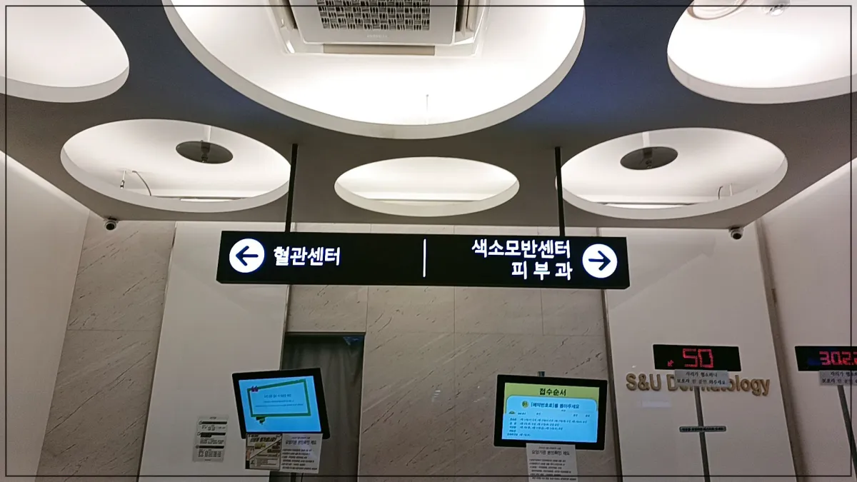 천장에-조명이-있고-가운데-천장형-에어콘보임-그아래-왼쪽-화살표로-혈관센터-오른쪽-화살표로-색소모반센터-피부과-라는-안내판-접수처