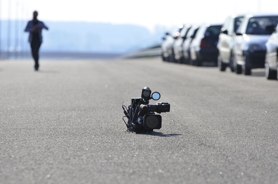 An abandoned video camera lies in the center of a road with parked cars and a distant person.