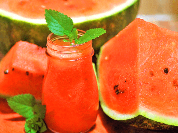 빨간색의 잘린 수박조각들이 있고 맨 앞에는 작은 유리병에 가득 담긴 빨간 수박주스가 있다.