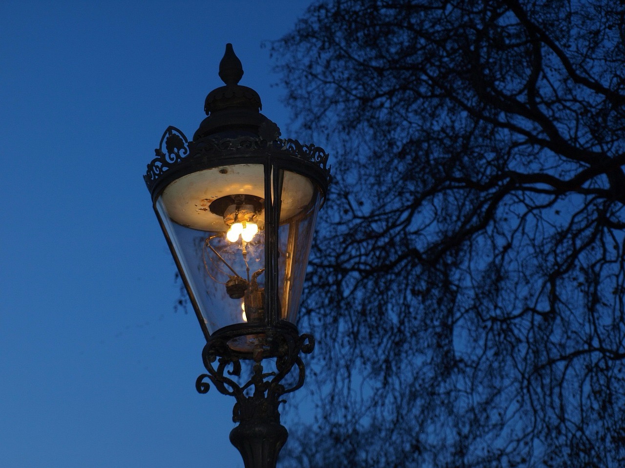런던의 고전적인 가로등이 어두운 저녁 하늘 아래 점등되어 있으며, 배경에는 음산한 분위기의 겨울 나뭇가지가 보인다