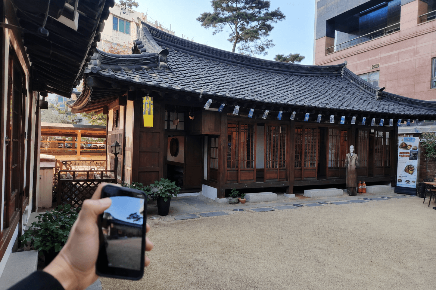 대구 중구 서상돈고택, 검은 기와지붕과 원목 기둥의 한옥 본채, 마당 앞 서상돈 선생 입상과 화분, 뒤편 현대 건물과 소나무가 보이는 대구 근대골목 전통 가옥 전경