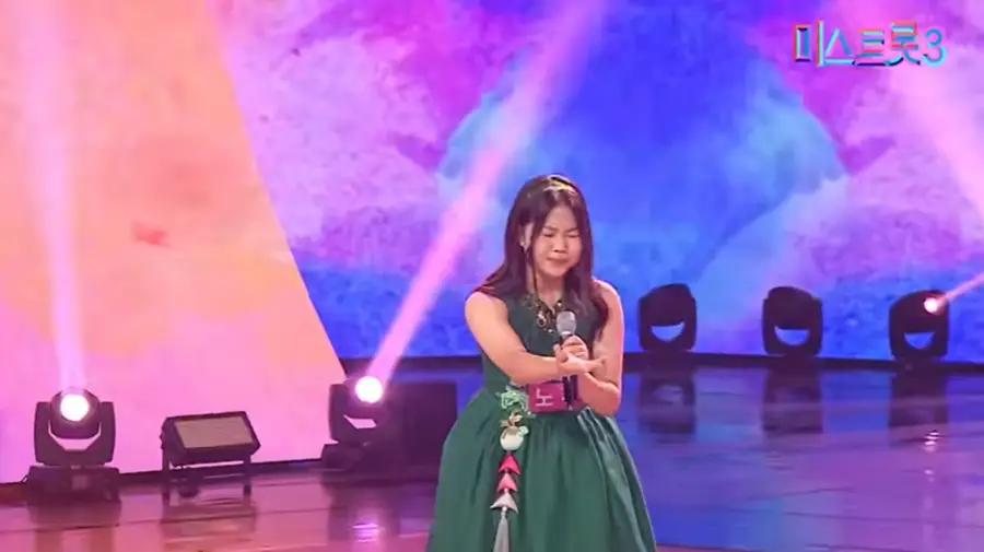 미스트롯3- 초록색 민소매 아래로는 퍼지는 치마를 입고 어깨까지 오는 머리에 금색 머리띠를 하고 왼손으로 마이크를 잡고 오른손은 먼곳을 가리키며 노래하는 노규리