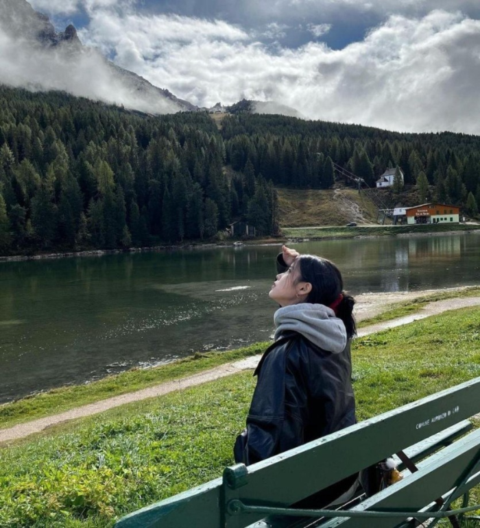 멋진 산맥과 작은 강을 앞에두고 벤치에 앉아 한손으로 햇빛을 가리며 하늘을 올려다보고 있는 아이유의 사진