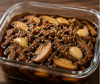 투명한 유리 용기 안에 윤기가 자르르 흐르는 갈색빛 소고기 장조림과 통통한 마늘, 버섯이 자작한 간장 국물에 폭 잠겨 통깨가 뿌려진 모습