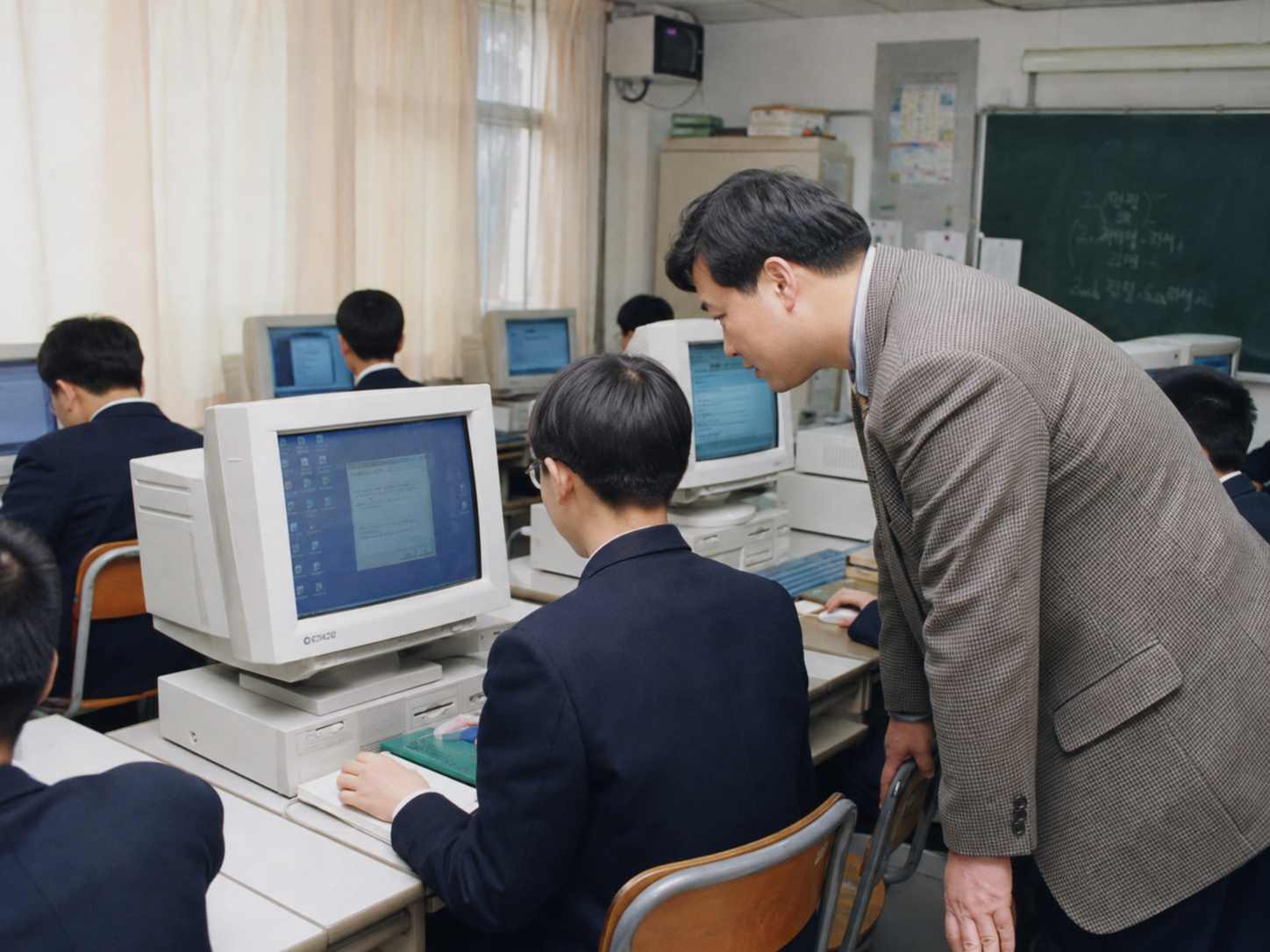 여러 대의 오래된 CRT 컴퓨터가 놓인 교실에서 학생들이 컴퓨터를 사용하고 교사가 수업을 돕는 모습