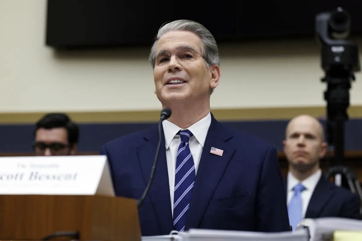 회색 머리의 백인 남성이 진한 남색 정장과 줄무늬 넥타이를 착용하고 청문회장 증인석에 앉아 있다. 그의 앞 테이블에는 'Scott Bessent'라고 적힌 명패가 보이며, 마이크가 설치되어 있다. 남성은 정면을 응시하며 약간 미소 짓고 있다. 배경은 베이지색 벽과 검은색 카메라 장비가 보이며, 뒤쪽에 다른 참석자들이 흐릿하게 보인다. 전형적인 미국 의회 청문회 현장의 모습이다.