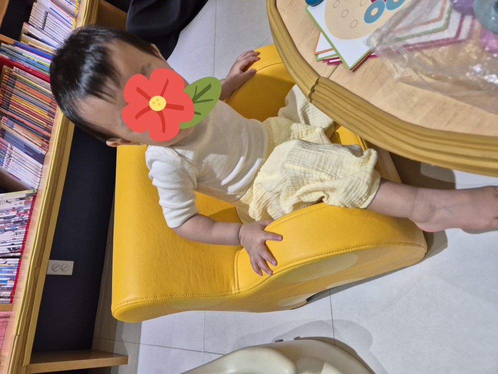 노란 소파 의자에 앉아 할머니의 설명을 들을 준비를 하고 있는 13개월 아기 모습