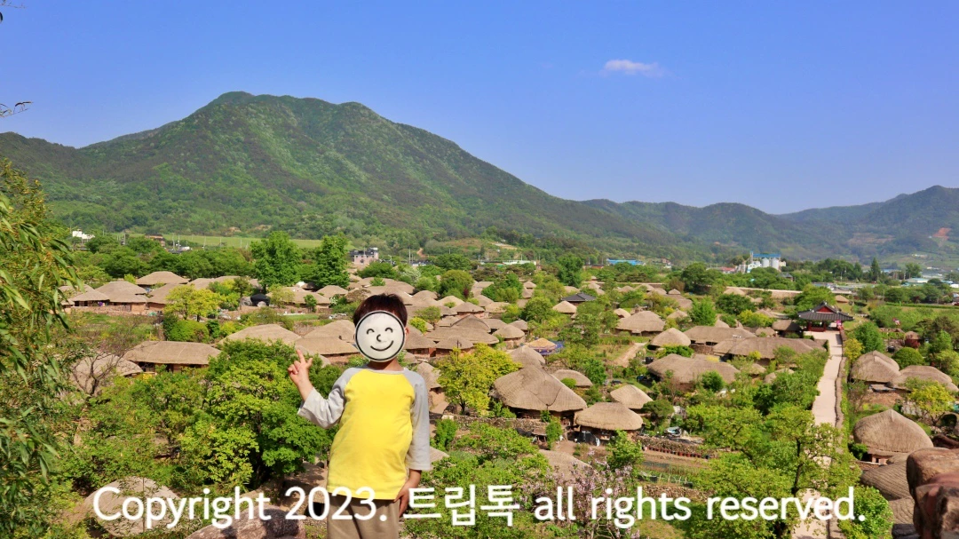 파란 하늘아래 버섯처럼생긴 초가지붕이 매우 많이 옹기 종기 모여있는 배경으로 노란옷을 입은 아이가 서있다