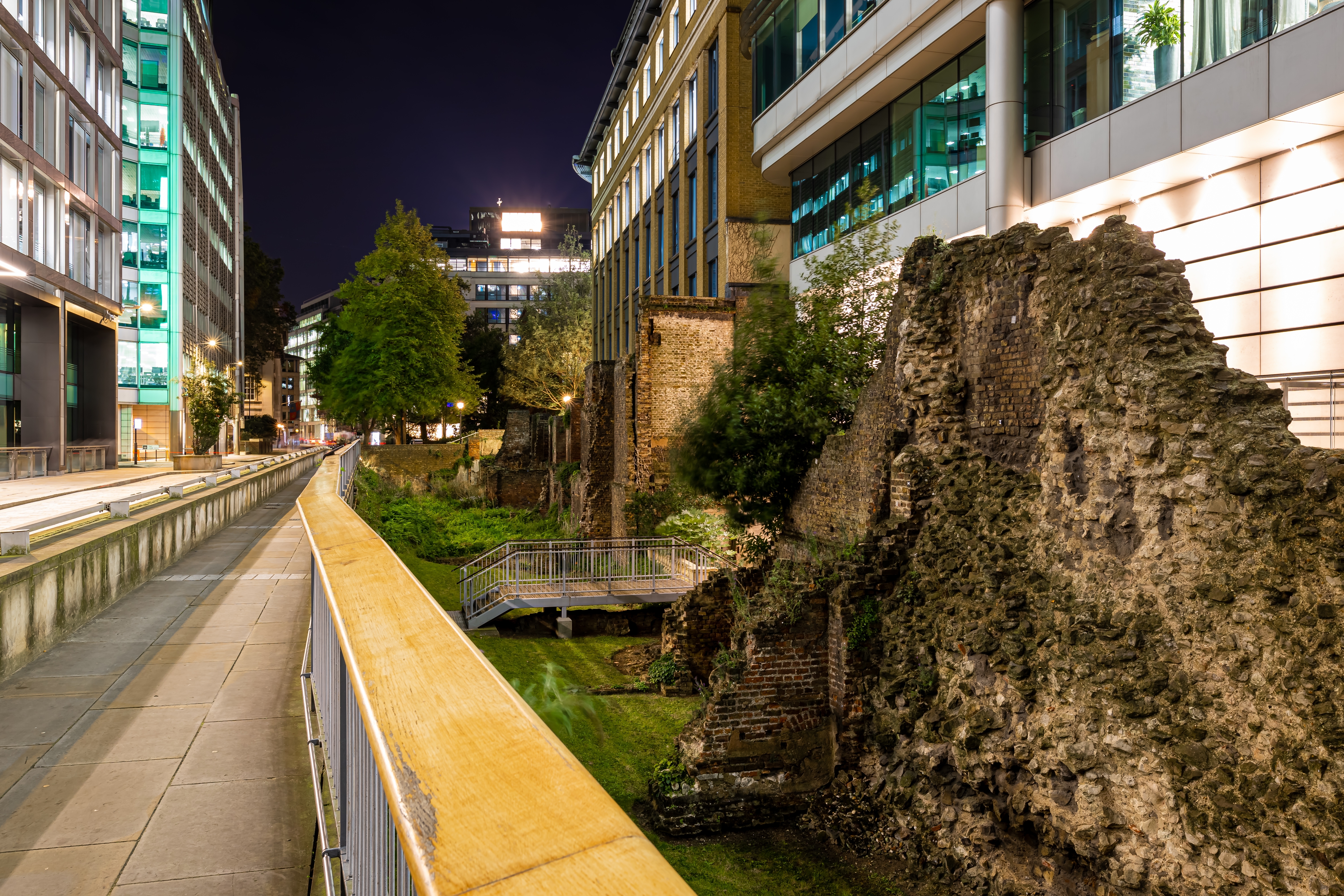 현대적인 건물 사이에 위치한 고대 로마시대 성벽인 런던 월(London Wall)의 야경 모습. 조명 아래 고풍스러운 석조 구조물이 도심 속에서 대비를 이루고 있음.