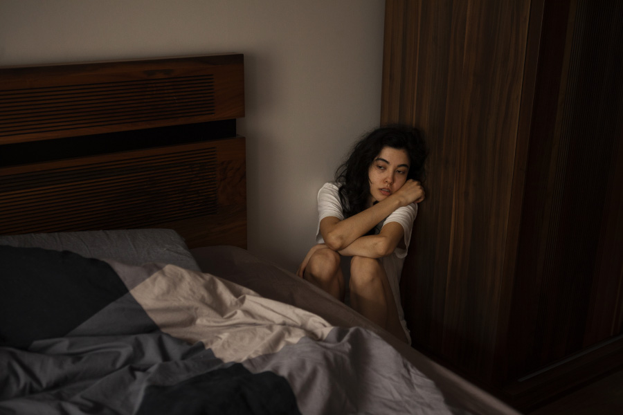 공황장애증이 있는 젊은 서양 여성이 침대 옆에 있는 벽의 모서리 부분에 쪼그리고 앉아서 불안, 초조감을 느끼고 있는 것을 찍은 사진