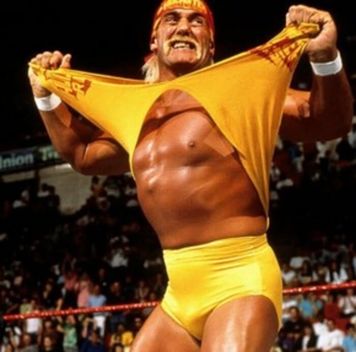 헐크 호건이 링 위에서 상징적인 노란색 셔츠를 찢으며 포효하는 장면. 관중석을 배경으로 전성기 시절의 에너지가 느껴진다.