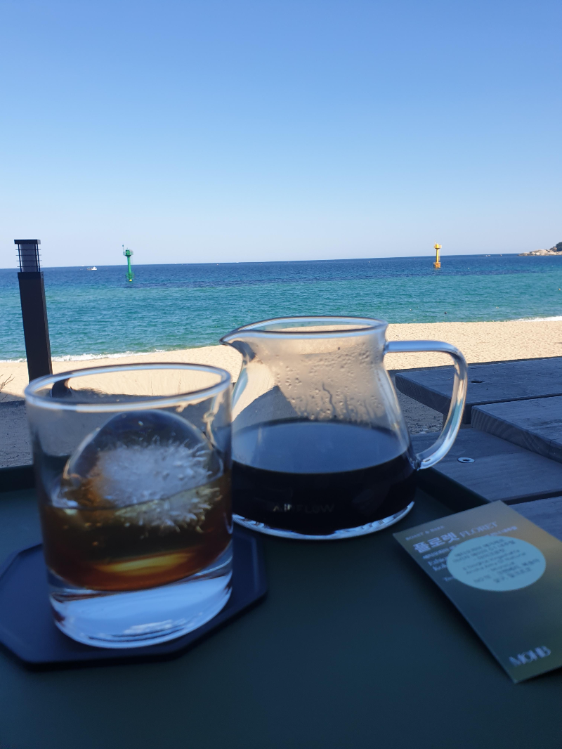 바다와 해변이 보이는 곳에 아이스 커피가 든 서버와 얼음이 든 유리컵이 테이블에 올려져 있다.