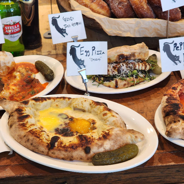 노릇하게 구운 조지아식 하차푸리 빵 위에 반숙 계란이 올려진 에그 피자, 옆에는 피클이 곁들여진 모습. 주변에는 다양한 피자와 샌드위치가 함께 진열된 빵집 내부