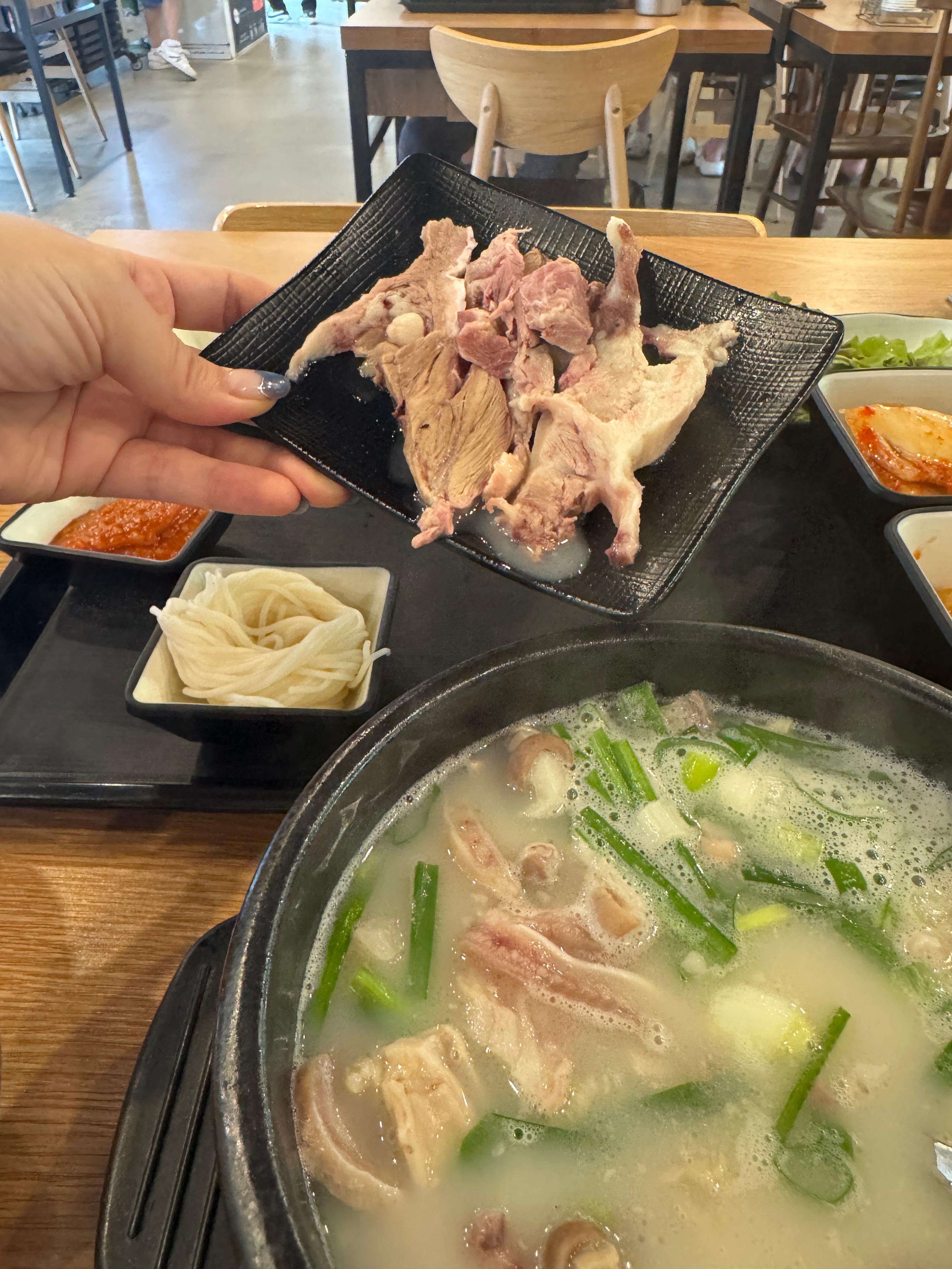 검은 접시에 담긴 수육을 손에 들고 있는 장면. 국밥 뚝배기와 소면&amp;#44; 김치&amp;#44; 쌈채소가 함께 보인다.