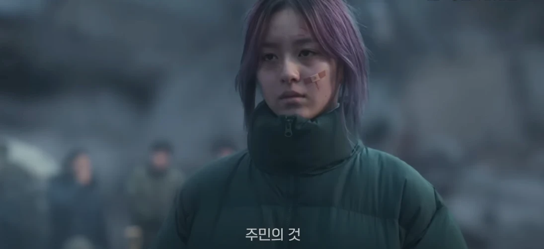 페허가 된 도시를 배경으로 초록색 패딩에 보라색 염색머리로 어딘가를 무표정하게 바라보고 있는 박지후