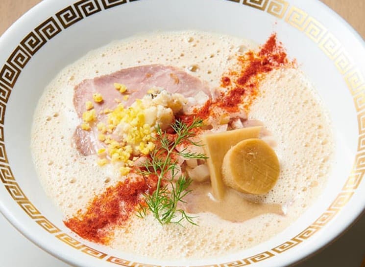 흰색 그릇안에 크림같은 국물이 있고 그 위로 차슈와 빨간색 가루가 올라가 있다